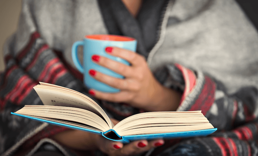 Photograph of a woman wrapped in woollies and a blanket, reading a book and holding a cuppa. Red nails.