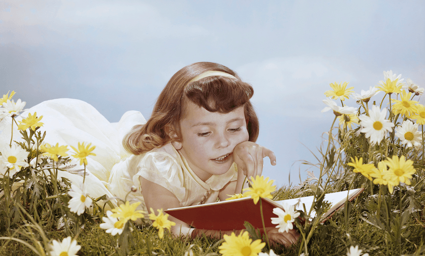 An old-timey photograph of a girl wearing a headband and lacey dress, lying in a dreamy meadow of daisies, reading a book. Bluebird sky.