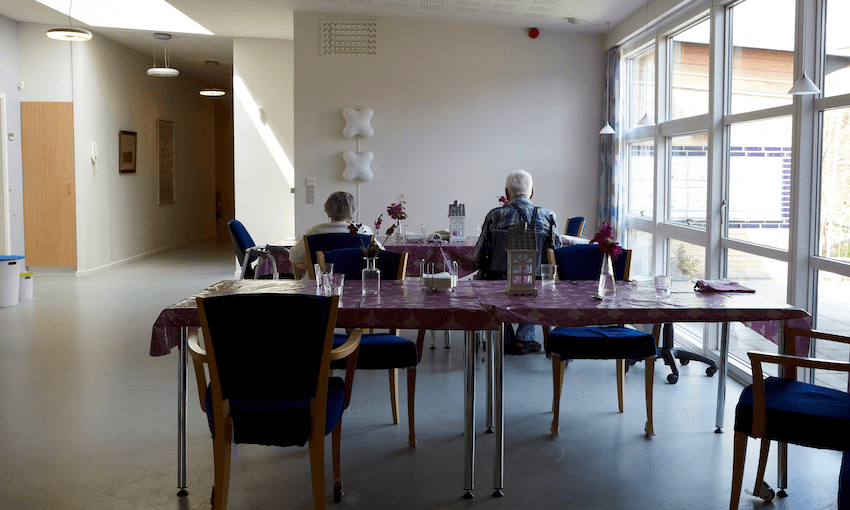 Interior of a rest home dining room; vinyl tablecloths, hard chairs, two residents seated.