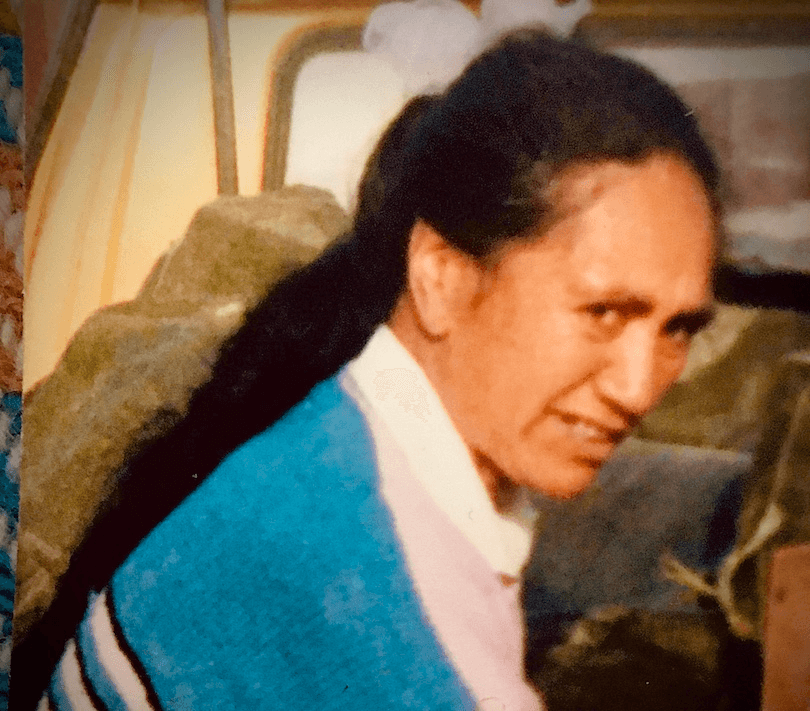 Old family photo of a middle-aged Māori woman, long hair in a ponytail, squinting and smiling at the camera.