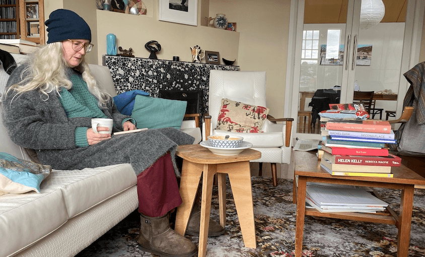 Writer Elizabeth Knox on a couch, wrapped up in a beanie and slippers, reading through a stack of books