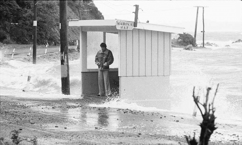 Man standing at a bus stop during a storm, Māhina Bay, Eastbourne (Photo: Evening post newspaper. Ref 1/4-022744-F. Alexander Turnbull Library) 
