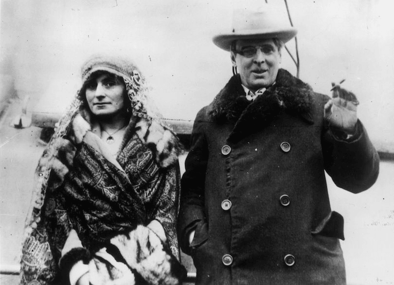 Old black and white photograph of a middle-aged couple walking toward camera, he is the centre of attention but she has a certain clever look in her eye