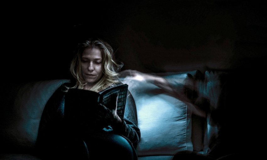 A dark room, a woman reads in bed, a ghostly arm reaching toward her