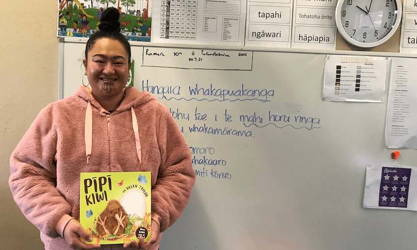 A Māori woman stands in front of a whiteboard, holding a picture book. Notes on whiteboard are in te reo. She's very proud of this space.