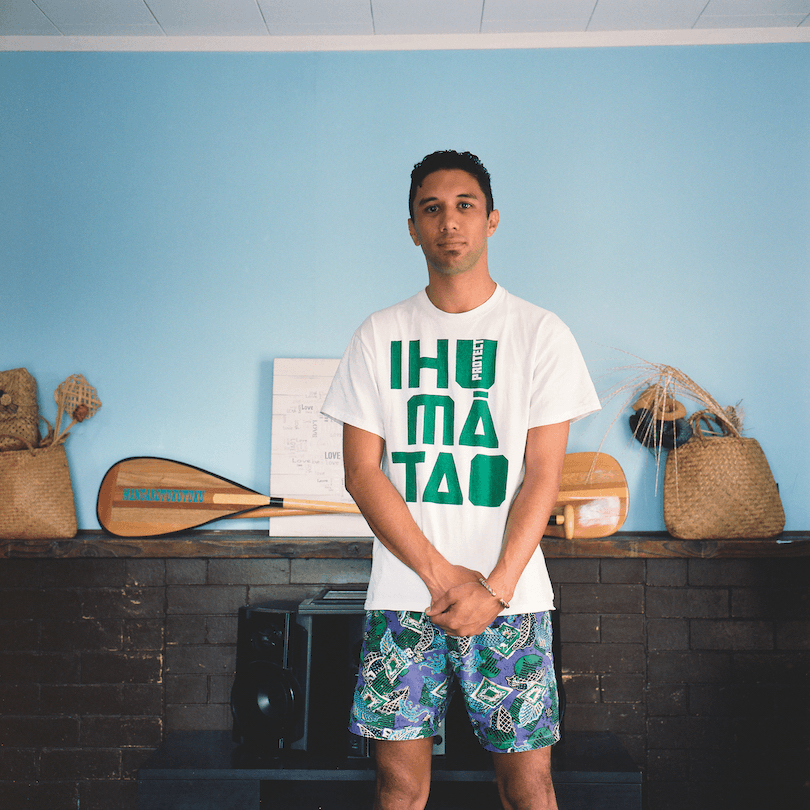 A young man wearing a Protect Ihumātao T shirt and boardies, stands in front of a mantelpiece display of kete and a waka paddle