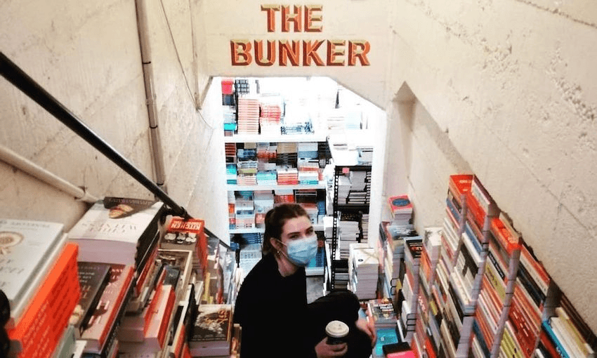 A young woman holds a takeaway coffee and perches on the steps down to "The Bunker", Unity's storage room, with glorious stacks of books all around.