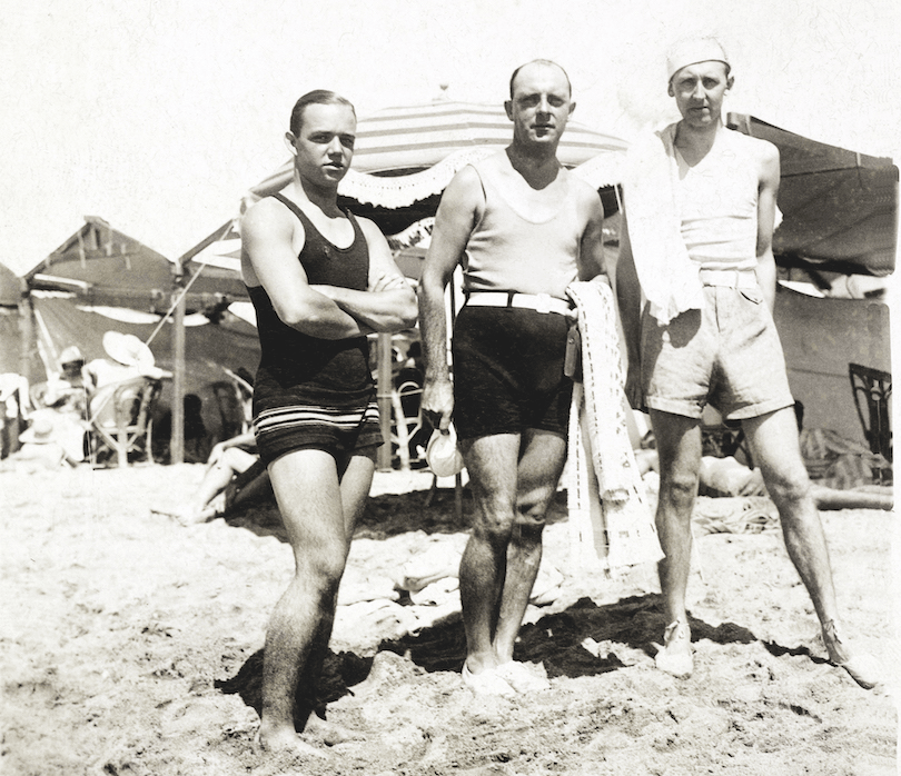 Black and white photo showing three men in front of what appear to be barracks. They're standing in sand.