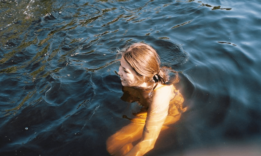 A young woman submerged in water up to her shoulders, dark water, outdoors, sun on her face, absolutely radiant