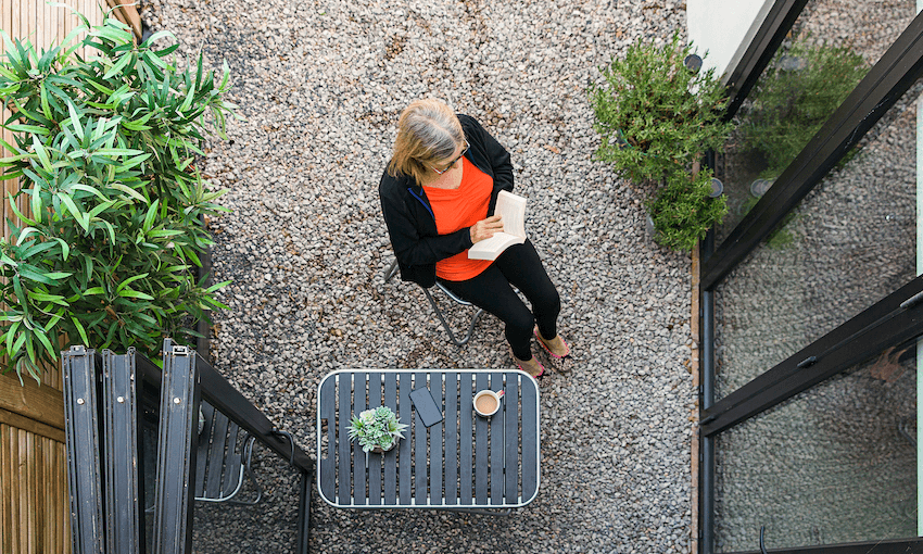 View from above of a senior woman reading in her courtyard, phone and coffee on a little table beside her. Lockdown vibe.