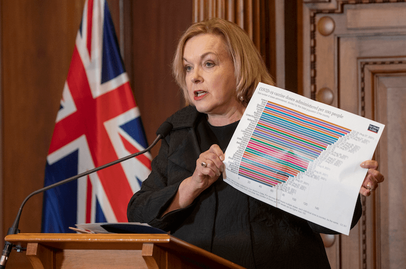 a white woman in her early 60s with blonde hair, standing at a podium talking with a graph in her hands 