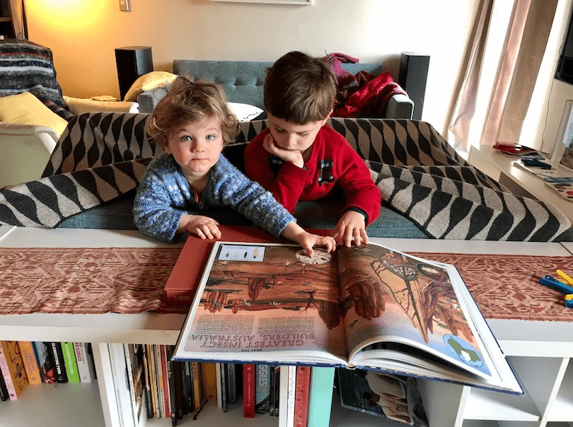 Two young kids in a blanket fort, poking their heads out to read a huge picture book. 