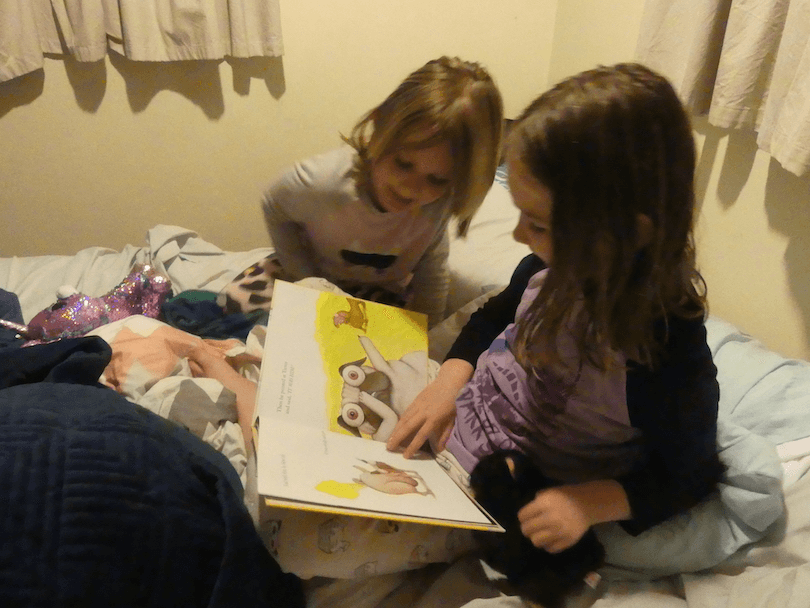 Two girls reading a picture book, sitting on a bed