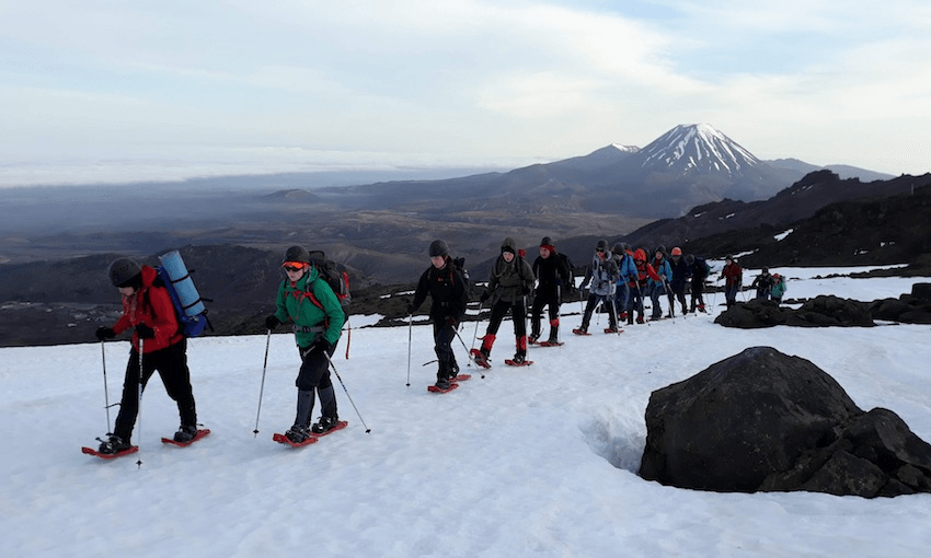 A trail of students hike through the snow, a snow-topped mountain can be seen in the background.