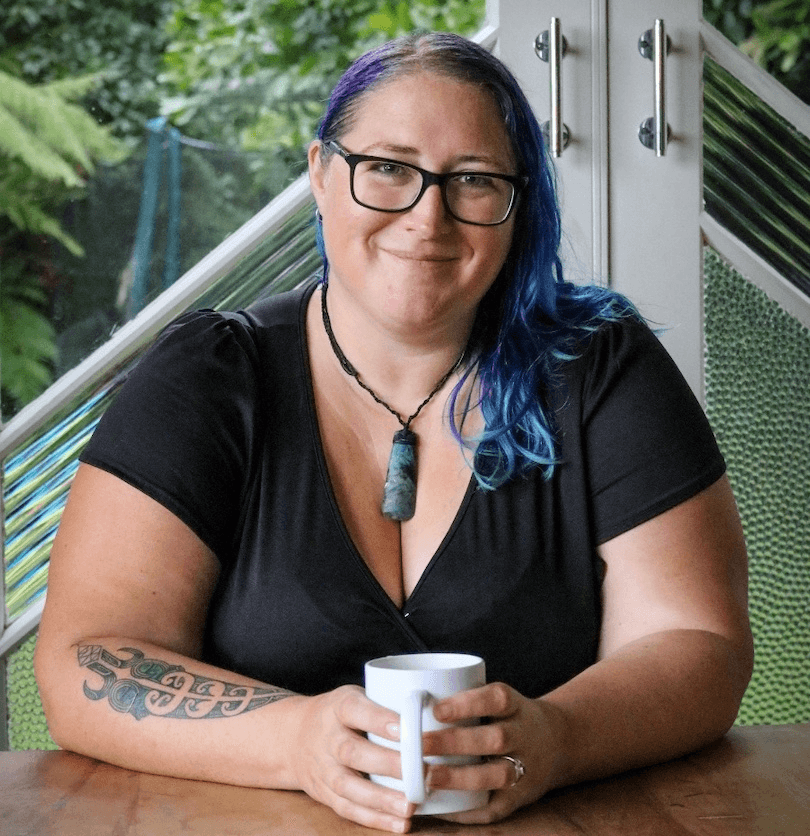 Smiling woman seated at a wooden table, hands wrapped round a cuppa, pounamu around her neck. 
