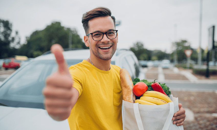 A young hipster wearing a bright yellow T and clutching a reusable bag of produce, in a carpark, giving a huge cheesy thumbs-up.