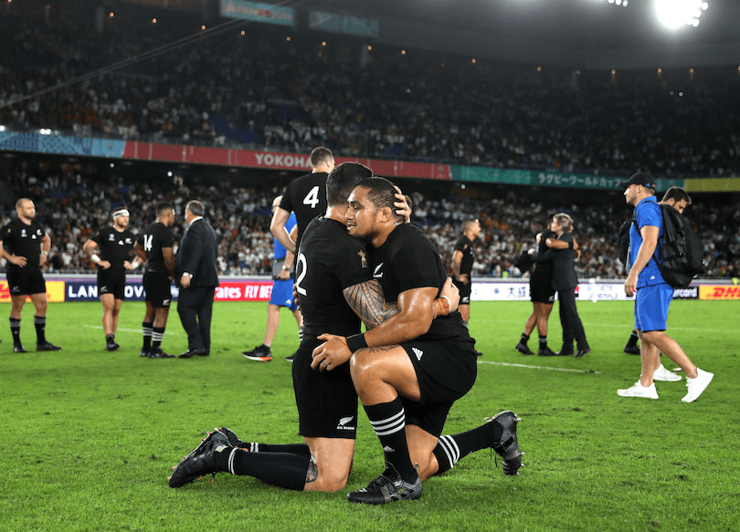 Sonny Bill Williams and Ofa Tu'ungafasi in the foreground, in All Blacks kit, embracing, on their knees; in the background, aftermath of a rugby match. 