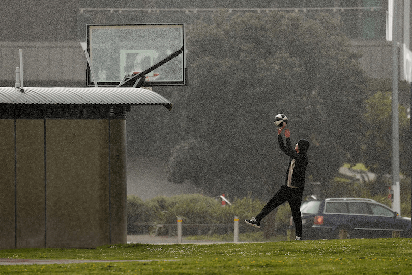 Photograph of a drizzly day, a teenager in a tracksuit shooting hoops by himself