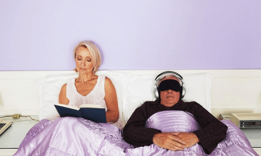 A couple in bed (their bedroom is garish lilac): the woman's sitting up happily reading a book, the man's hunkered down in an eye mask and headphones.