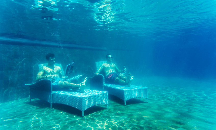 Two men on deckchairs at the bottom of a pool, one is reading, one gazing off into the distance.
