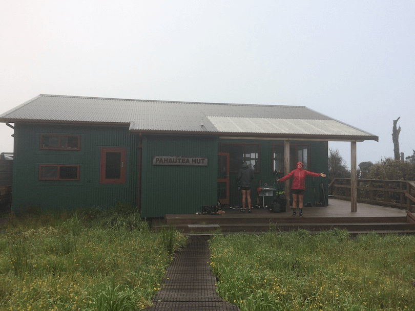 Exterior shot of a fancy-as DOC hut, a tramper standing triumphantly on the porch. Grey drizzly day. 