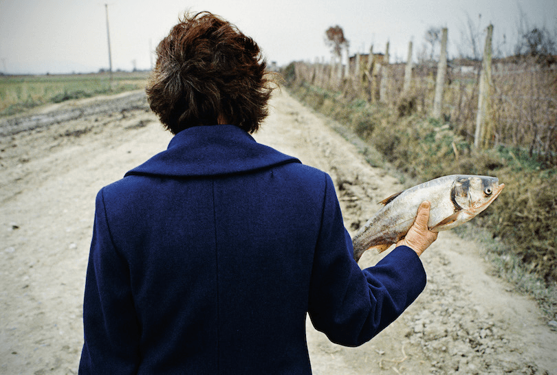 Odd photo of a woman holding a fish at shoulder height, pointing away from her body. She's walking on a gravel road, she has a coat on, we're watching her from behind. 