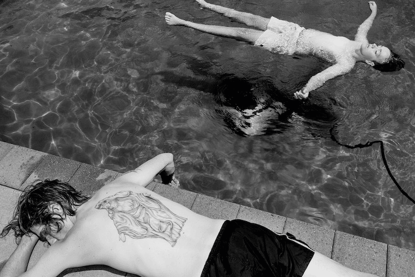 Black and white photograph: a child floats in a swimming pool, face-up, blissed out; a person lies face-down on the side of the pool, one hand dangling in the water. 