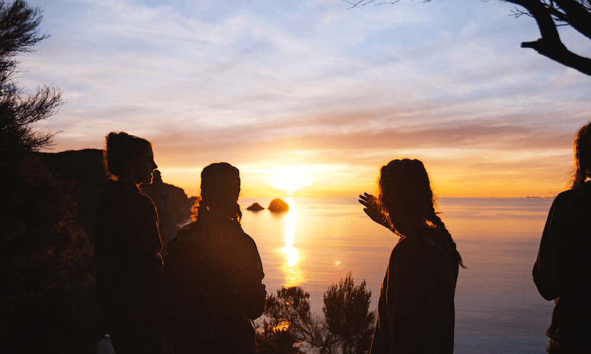 Three students look out across the ocean from a hilltop, silhouetted by the setting sun.