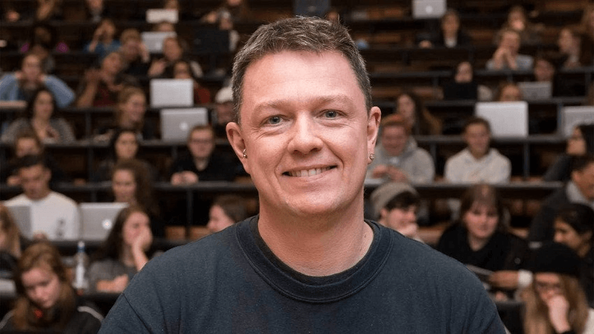 a white man with a slight smaile and brown hair wearing a charcoal coloured tshird in front of a lecture theatre of students with ubiquitous macbook airs