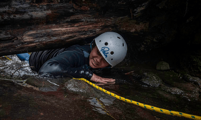 A student wearing a helmet crawls through a small gap in a rock, grinning and holding a rope.