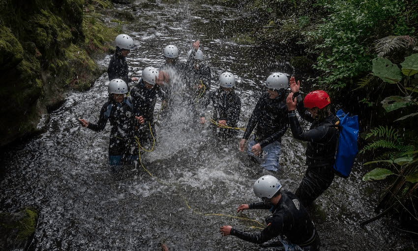 A group of students play in a stream, wearing helmets and wetsuits.