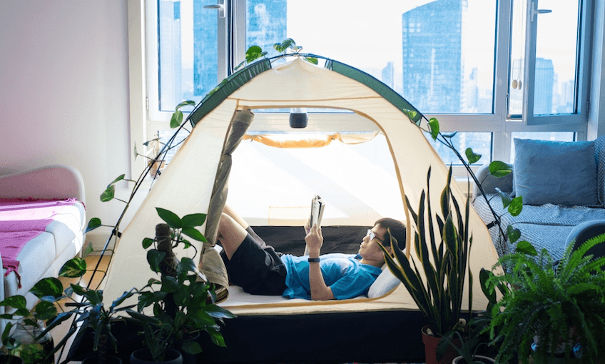A man has set up a pup tent in the lounge of his apartment, arranged all his houseplants around it and is lying down reading inside. Weird lockdown vibe.