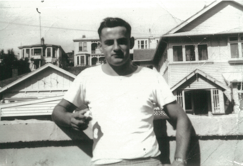 Black and white photo of a young man in a white Hanes T, leaning on a fence. Houses and hot sky in background.