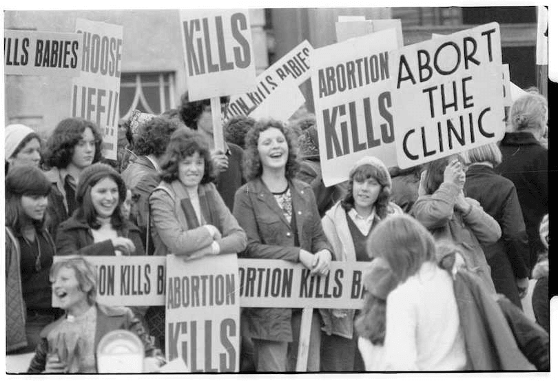 Black and white photograph of a crowd of protestors holding anti-abortion signs. Many are young women, grinning.