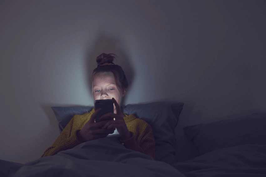A young teenager is sitting in her bed in the dark with only the light of her phone shining on her face.