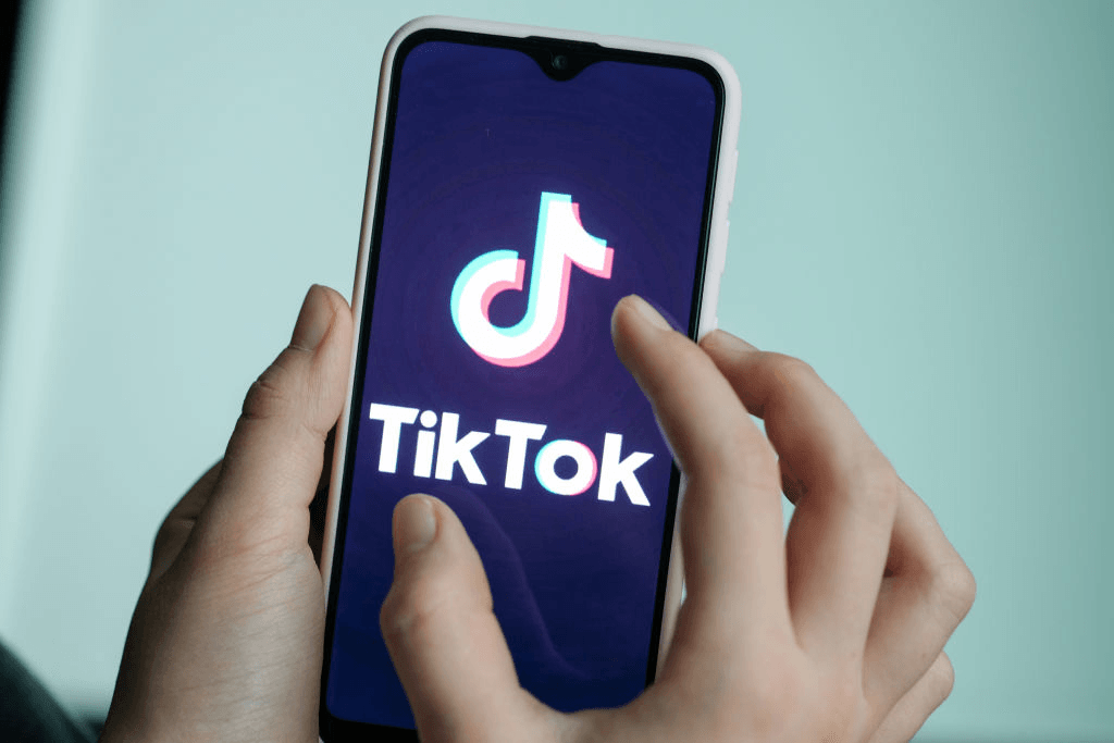 grey blue background, a hand with medium tone skin holding up a phone lit up with the tik tok icon