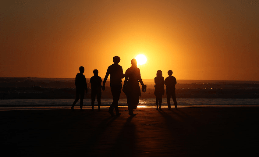 An image split in two: orange sunset sky, black wet sand. Six silhouettes of people, all in pairs, face the water and the setting sun.