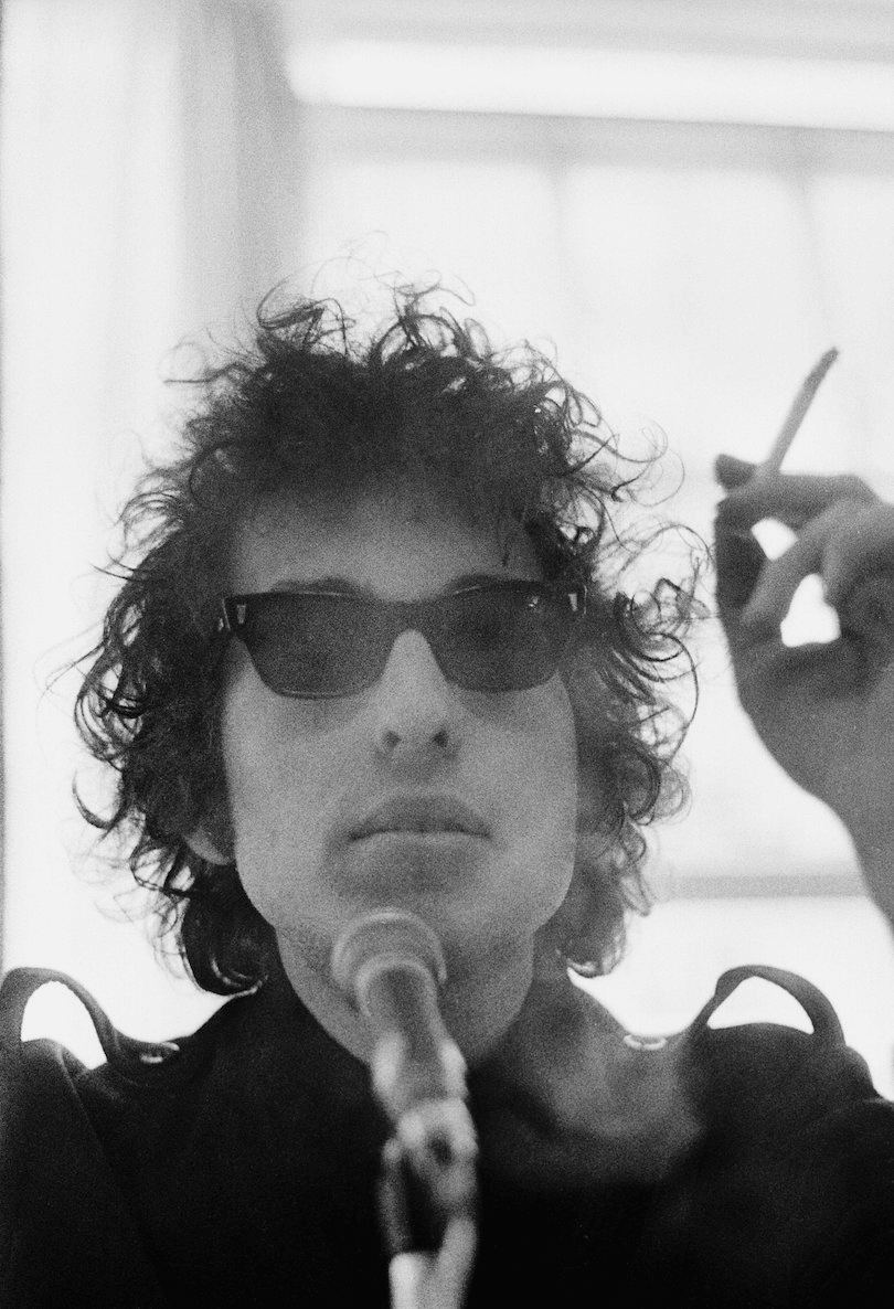 Black and white head and shoulders photo of middle-aged man, wild dark hair, shades, a microphone in his face and a smoke in his hand.