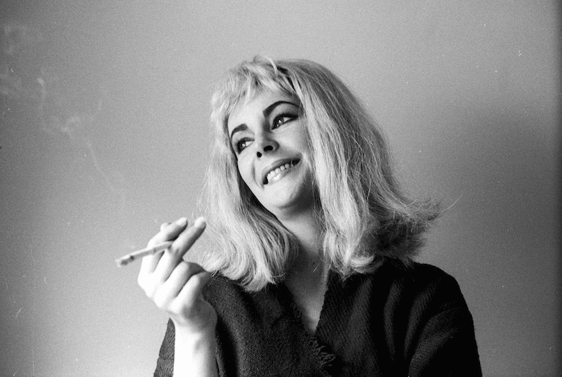Elizabeth Taylor in a blonde bob wig, photographed against bare wall, holding cigarette, smiling.