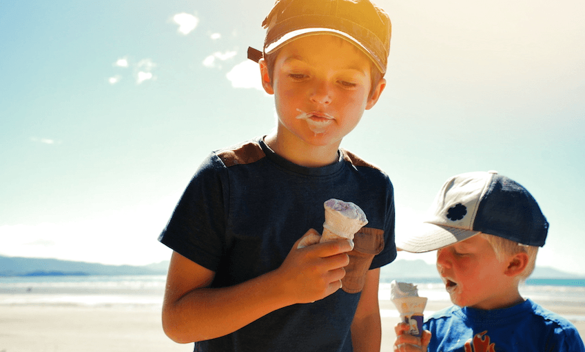 Two children with caps and t shirts on, ice creams in hand, concentrating pretty hard on them. Beauty of a day at the beach.