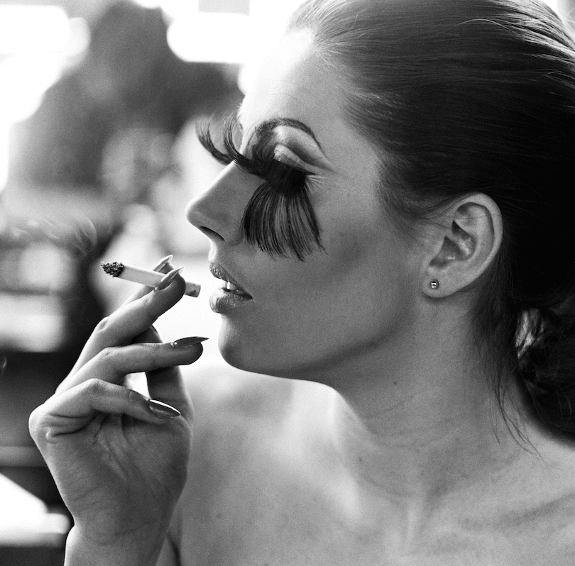 Black and white photo showing the profile of a young woman wearing ludicrously long false lashes – they reach her eyebrows and brush all the way down to the base of her nose. Smoking a cigarette, loving it. 