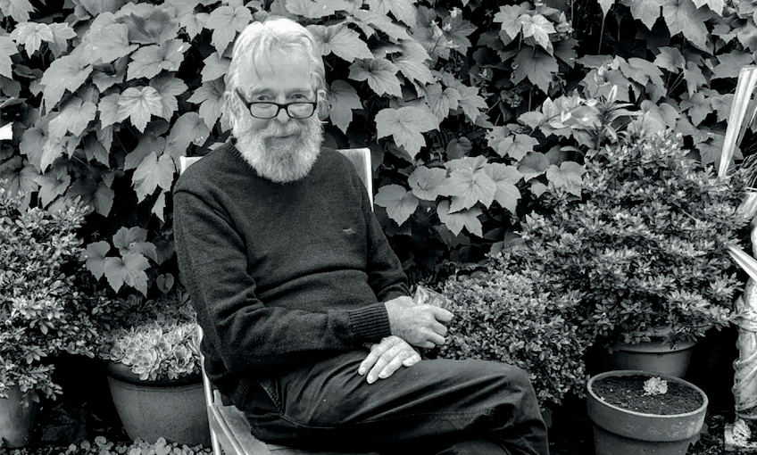 Black and white photograph: an older man, slight frame, switched-on look, seated in a garden. Legs crossed, he's holding a drink and looking to camera.