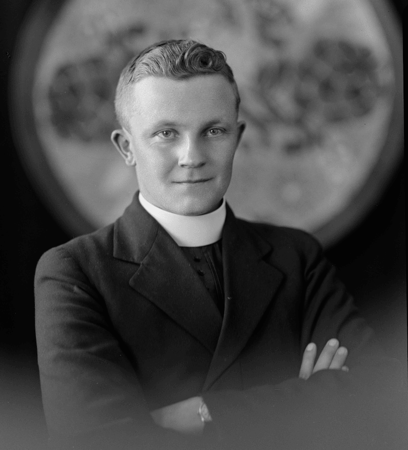 Photograph of a young, dapper priest, arms folded, looking confident and keen.