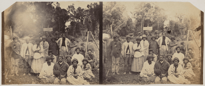 Sepia-toned photograph of a group of approximately 25 Māori gathered close together, some seated, most facing the camera. They're outdoors – beautiful bush in the background.