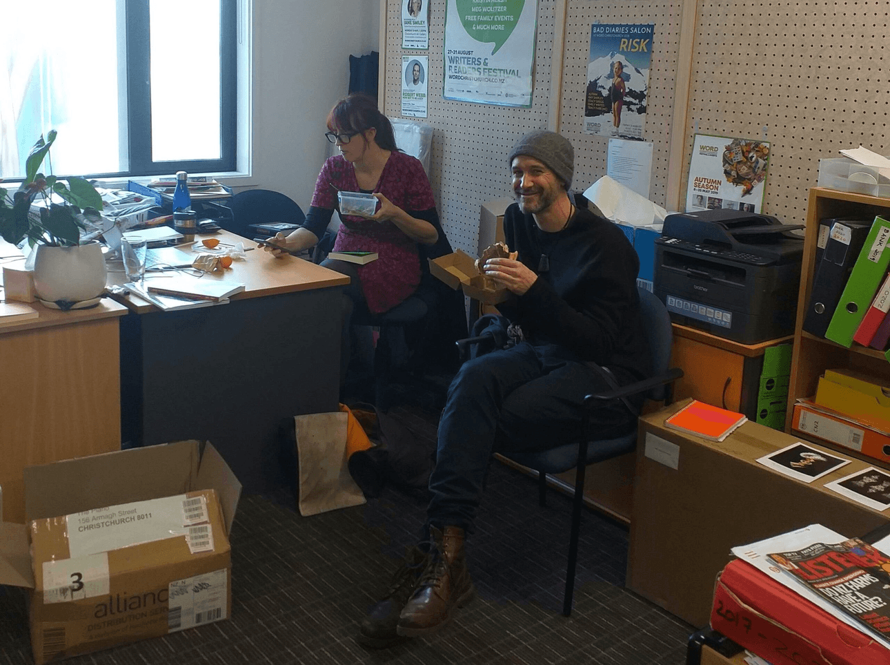 A thrown-together office room, boxes and bits and pieces everywhere, two people eating lunch. 