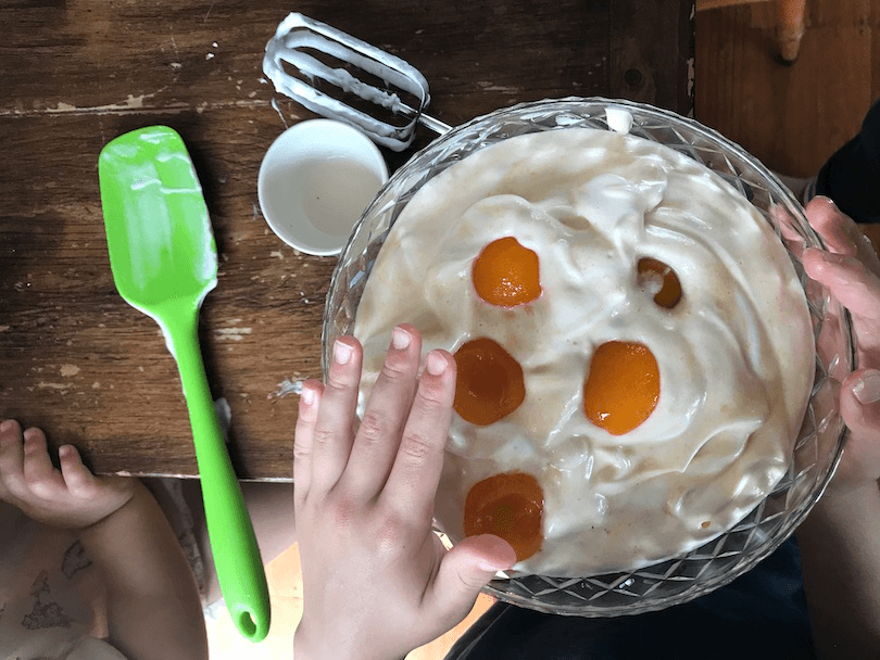 A bowl of white frothy-looking stuff with canned apricots pocked on top. We can the hands of two children who are clearly interested in getting a nosy at this monstrosity.