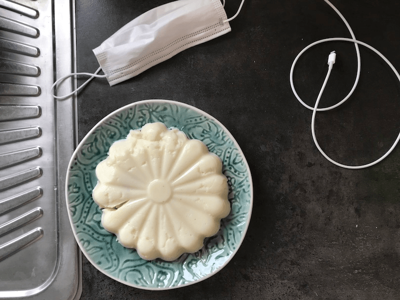 Aerial view of a kitchen bench featuring phone charger, disposable mask, and a pretty little white pudding, set in the shape of a flower. 