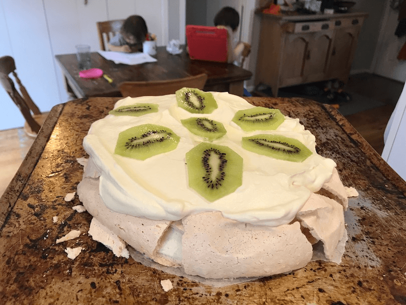Background: kids drawing and on iPad at dining table. Foreground: a very flat pavlova, with cream and kiwifruit. 