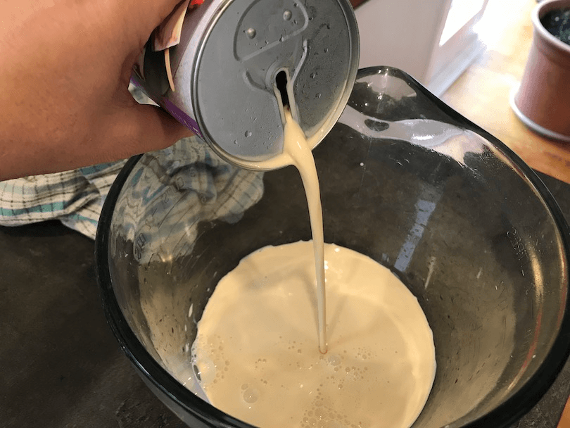 Pouring a can of evaporated milk into glass jug. Notably, the opening on the can is a long slit, rounded at top and bottom.