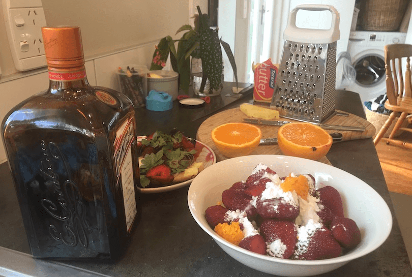 Kitchen bench strewn with stuff, including Cointreau, cut oranges, strawberry tops, spoons etc, a grater. Foreground: a bowl of strawberries covered in icing sugar and orange rind.
